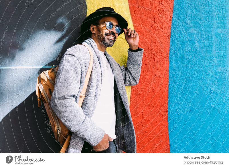 Stilvoller hispanischer Mann, der sich an eine Graffiti-Wand lehnt und seinen Hut berührt Lächeln Streetstyle positiv berühren urban Straße Gebäude männlich