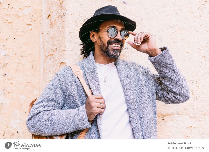 Fröhlicher hispanischer Mann, der seine Sonnenbrille zurechtrückt Streetstyle ausrichten Lächeln Wand bröckeln Straße Glück urban männlich berühren Hut Rucksack