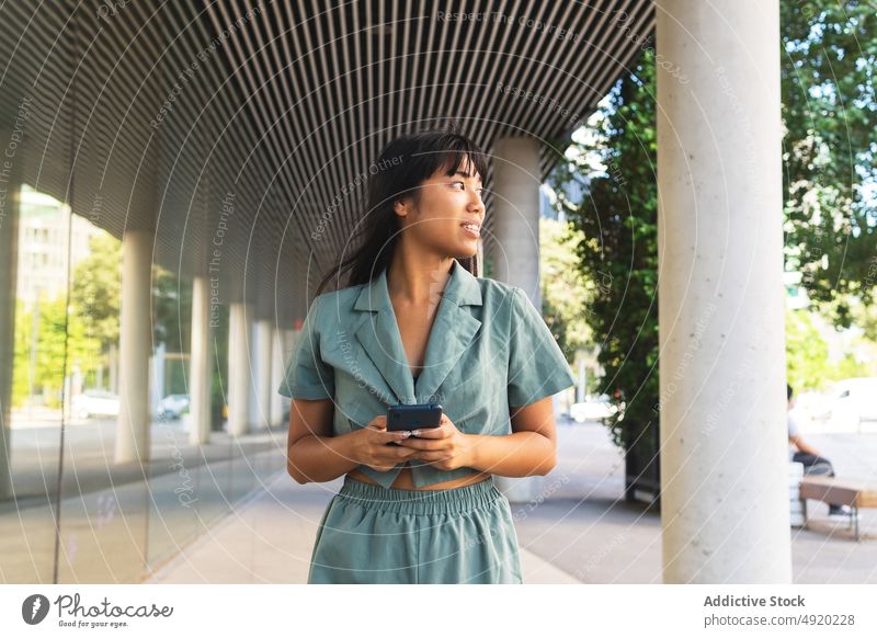 Fröhliche asiatische Frau benutzt Smartphone in der Nähe einer Glaswand benutzend Lächeln Texten Straße Sommer Stil Glück jung ethnisch positiv heiter Mobile