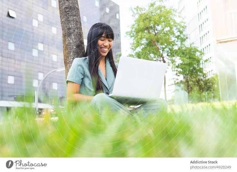 Studentin benutzt Laptop in der Nähe eines Baumes Frau Schüler heiter benutzend Hausaufgabe Rasen Bildung froh Park lernen Glück jung Sommer Gerät Lächeln