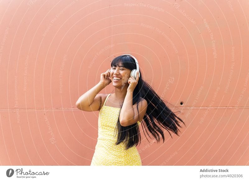 Glückliche asiatische Frau beim Musikhören Tanzen Lächeln zuhören Wand urban Kopfhörer berühren meloman jung ethnisch Straße Stil Klang Audio Gerät Gesang froh