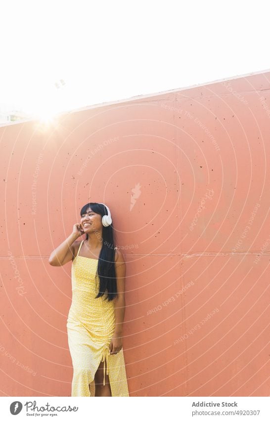 Glückliche asiatische Frau beim Musikhören Lächeln zuhören Wand urban Kopfhörer berühren meloman jung ethnisch Straße Stil Klang Audio Gerät Gesang froh