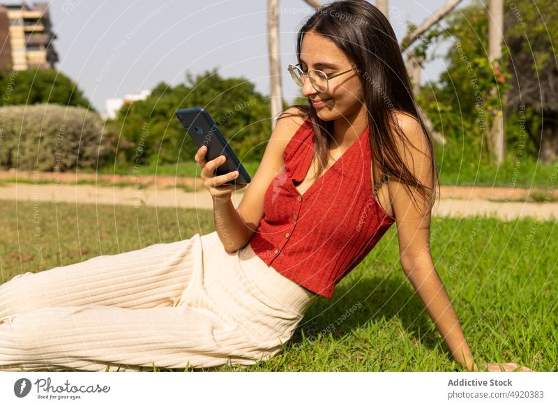 Lächelnde Frau, die auf einer Wiese auf ihrem Smartphone surft Rasen ruhen Textnachricht Park Browsen online soziale Netzwerke Talkrunde App Kälte Freizeit