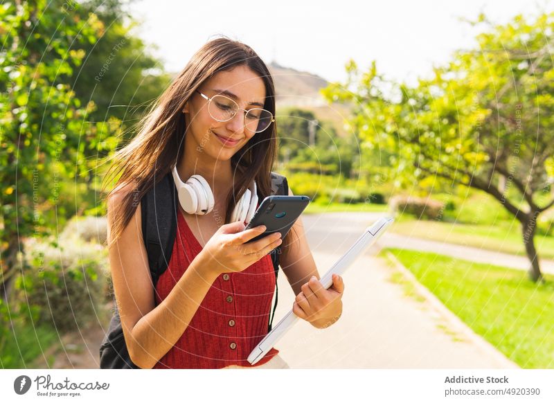 Fröhlicher Student mit Laptop, der im Park auf seinem Smartphone surft Frau Textnachricht Schüler Browsen online lernen soziale Netzwerke App Baum heiter