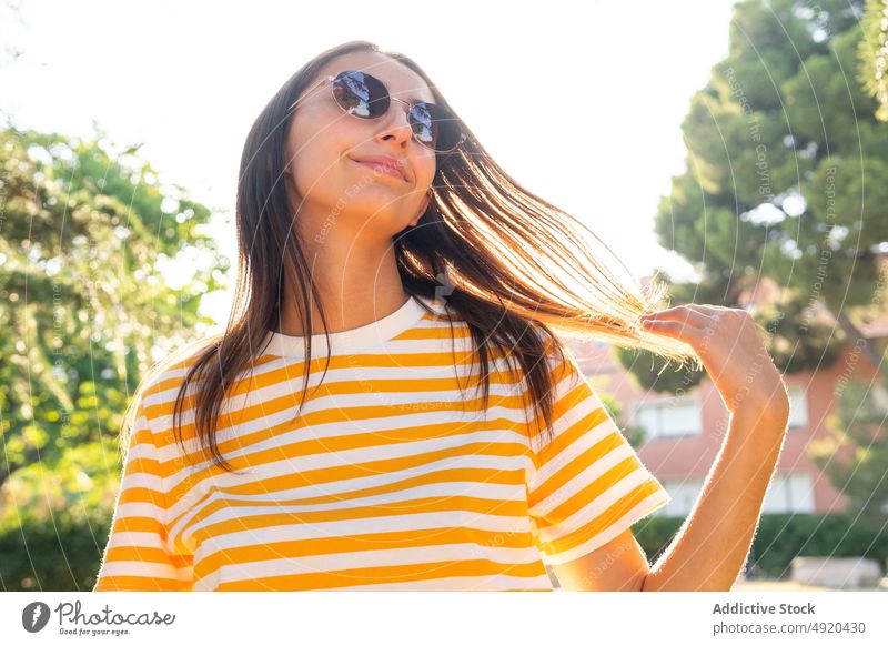 Junge Frau berührt Haare im Park Lächeln Haare berühren Sommer ruhen Wochenende tagsüber sonnig Glück dunkles Haar jung Stil Optimist Sonnenbrille froh positiv