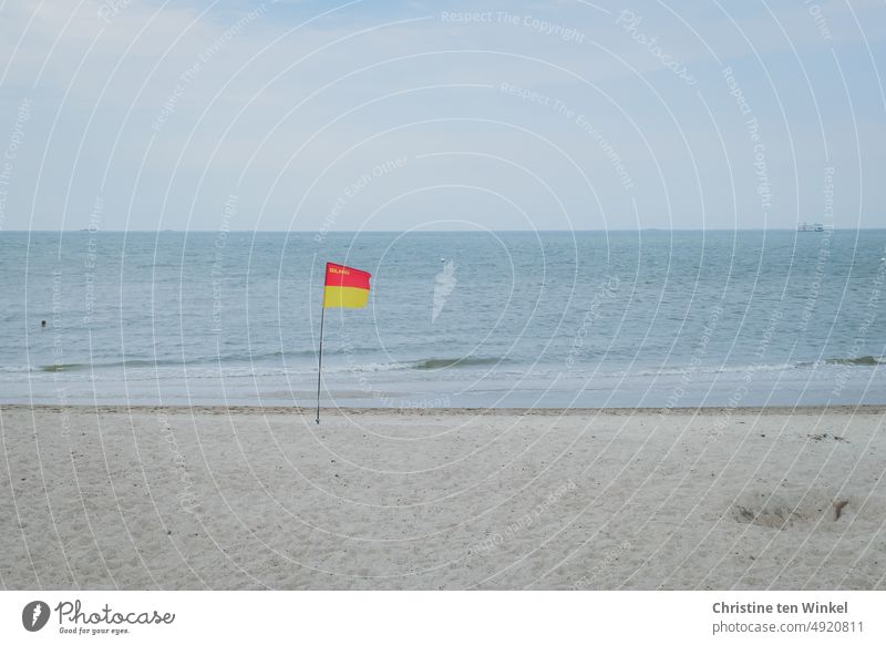 Eine DLRG Flagge weht bei trübem Wetter am leeren Nordsseestrand, im Wasser ist der Kopf eines Schwimmers zu sehen. Wasserrettungsorganisation