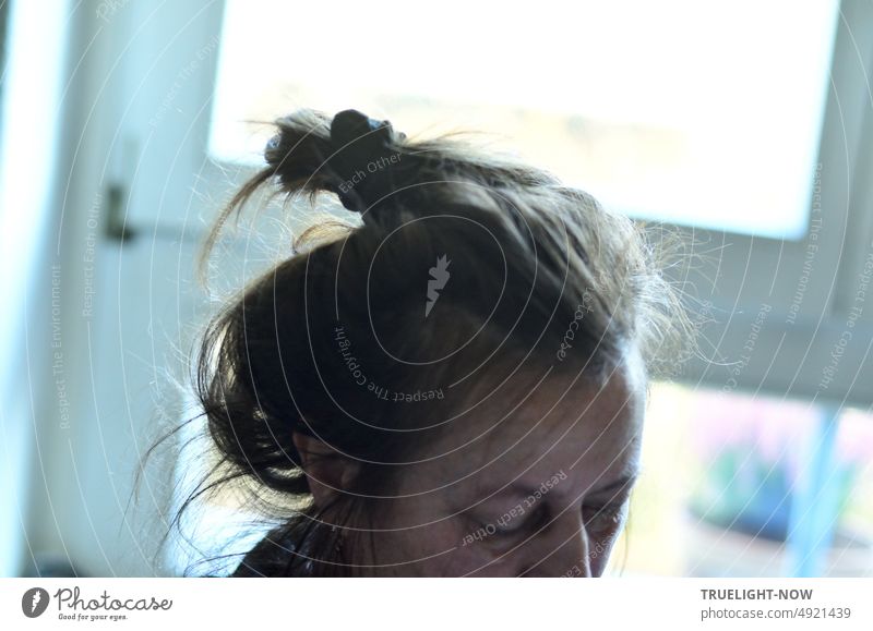 Leicht nach unten geneigter Frauenkopf Teilansicht mit stylish lässig gebändigter Haarpracht am Fenster im Gegenlicht konzentriert beschäftigt Kopf Haare