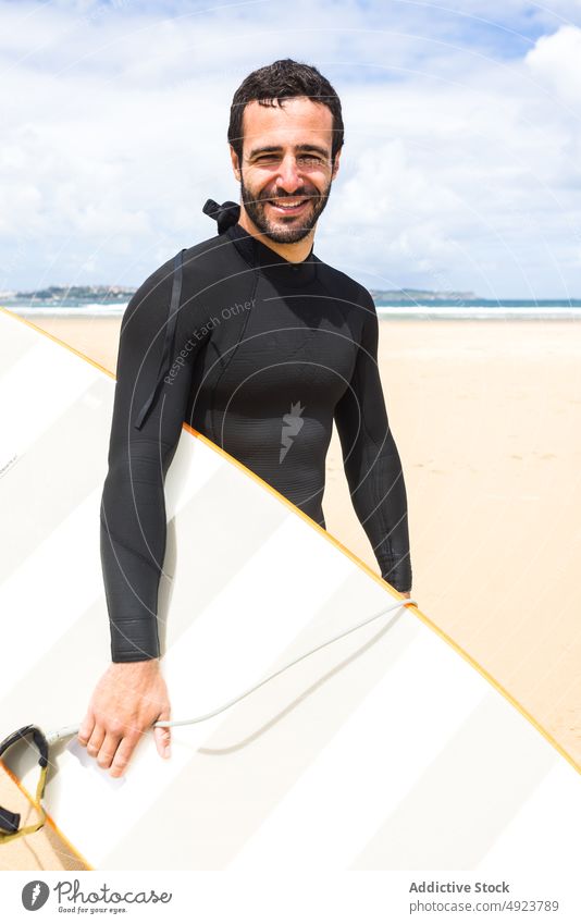 Glücklicher junger ethnischer Mann hält lächelnd ein Surfbrett am Meeresufer Surfer Strand Lächeln Shaka Athlet selbstbewusst hawaiianisch lose hängen männlich