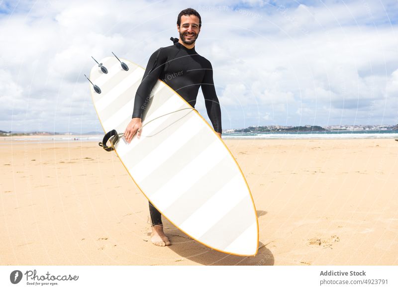 Glücklicher junger ethnischer Mann hält lächelnd ein Surfbrett am Meeresufer Surfer Strand Lächeln Shaka Athlet selbstbewusst hawaiianisch lose hängen männlich