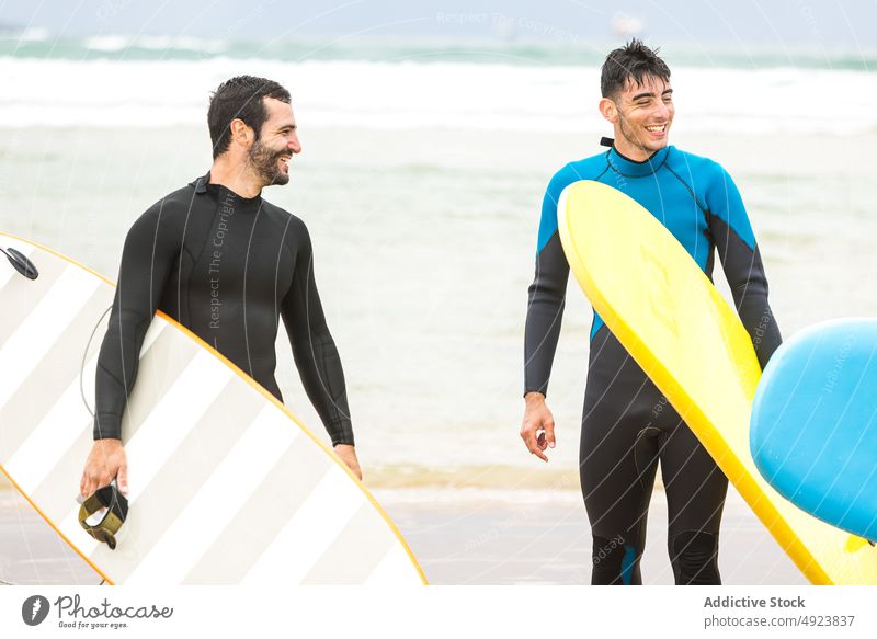 Athletische ethnische männliche Surfer mit Brettern, die gemeinsam am sandigen Meeresufer spazieren gehen Männer Spaziergang Strand Surfbrett Sport Freund