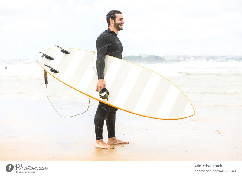Glücklicher junger ethnischer Mann hält lächelnd ein Surfbrett am Meeresufer Surfer Strand Lächeln Athlet selbstbewusst hawaiianisch lose hängen männlich Halt