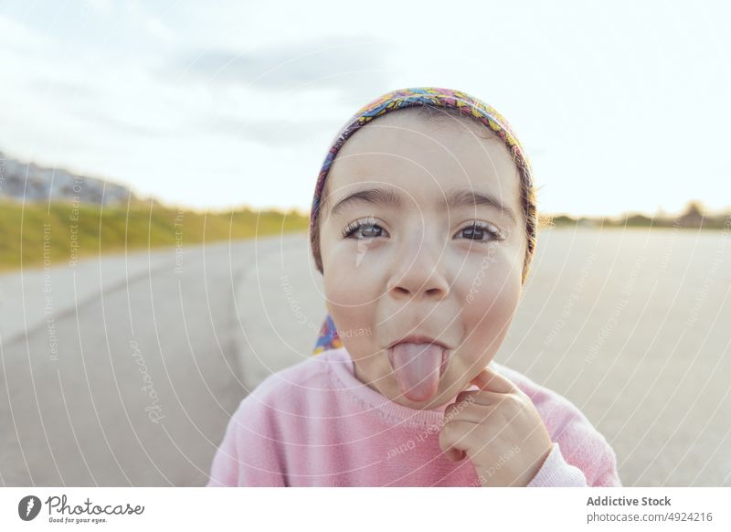 Lustiges hispanisches Mädchen zeigt Zunge Zunge zeigen Glück Grimasse lustig spielerisch Straße Wochenende wolkig niedlich Dummerchen Kind Asphalt heiter