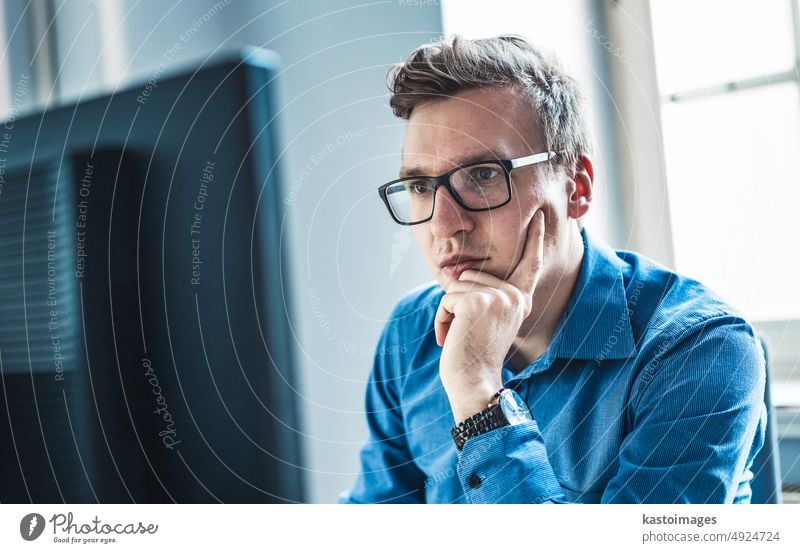 Hübscher junger Geschäftsmann mit Brille, der an seinem Tisch im Büro sitzt und den Bericht auf seinem Computerbildschirm betrachtet Monitor Mann Porträt
