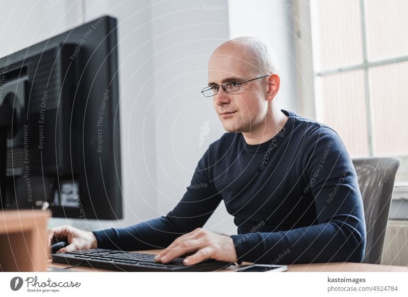 Hübscher junger Geschäftsmann mit Brille, der an seinem Tisch im Büro sitzt und den Bericht auf seinem Computerbildschirm betrachtet Monitor Mann Porträt