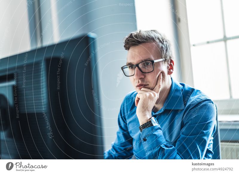Hübscher junger Geschäftsmann mit Brille, der an seinem Tisch im Büro sitzt und den Bericht auf seinem Computerbildschirm betrachtet. Monitor Mann Porträt