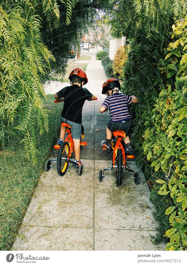 Zwei kleine Brüder fahren mit ihren Fahrrädern mit Stützrädern und Helmen auf dem Bürgersteig eines Vorortes Zwei Jungen wenig Vorstadt Viertel
