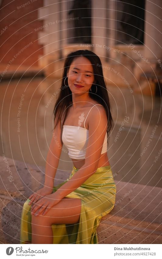 Schöne junge asiatische Frau sitzt in der Nähe von Brunnen in europäischen historischen Stadt in der Nacht mit Straßenlaternen auf. Weiblicher Tourist aus China auf Reisen in Europa. Mode und Stil Konzept.