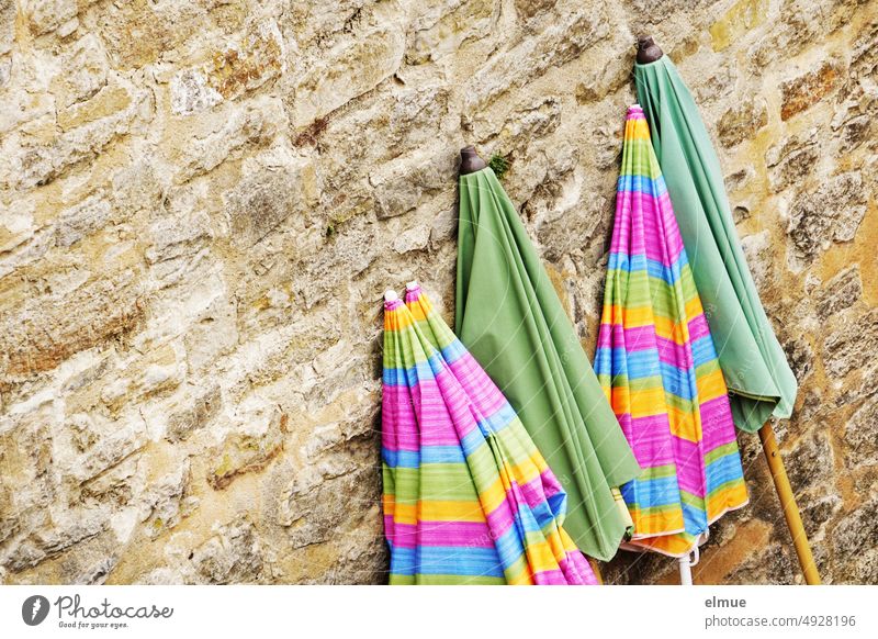 zwei grüne und drei farbig gestreifte, geschlossene Sonnenschirme lehnen an einer alten Steinmauer / Saisonende Sommer Schattenspender Steinwand Strandparty