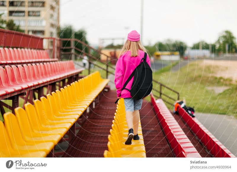 Ein Teenager-Mädchen mit einem Rucksack läuft durch die Tribüne des Schulstadions. Rückansicht Schule Schüler Schulzeit Bildung Frau Porträt Stadion Hintergrund