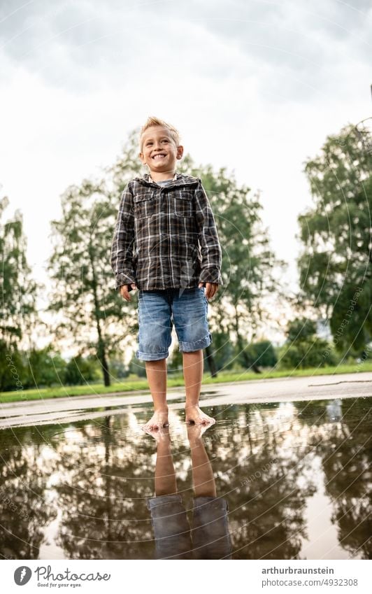 Junge in kurzen Jeans und Jacke steht barfuß in einer Regenpfütze am Spielplatz Wasser Wasseroberfläche Pfütze Regenwasser Kind Kindheit kindlich Spiegelung