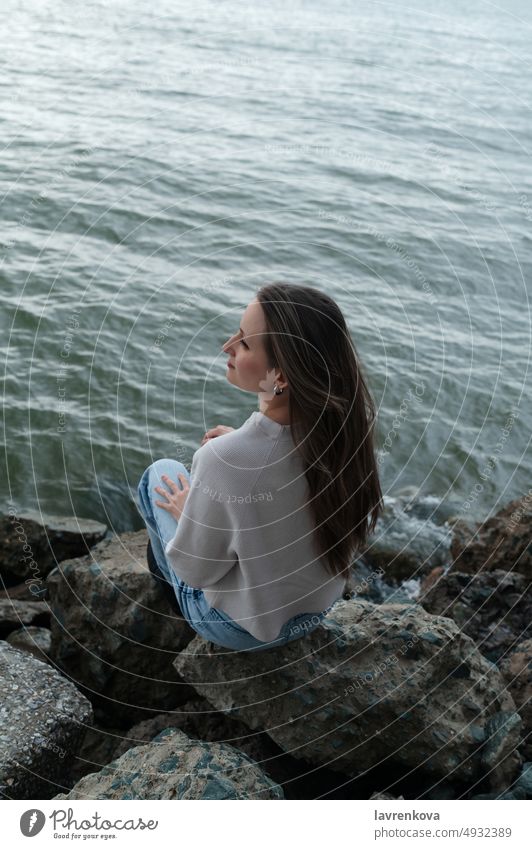 Junge Frau sitzt auf den Felsen am wilden Strand bei schlechtem Wetter Behaarung entspannend Erholung jung im Freien MEER Ufer Sitzen Küstenlinie Hafengebiet