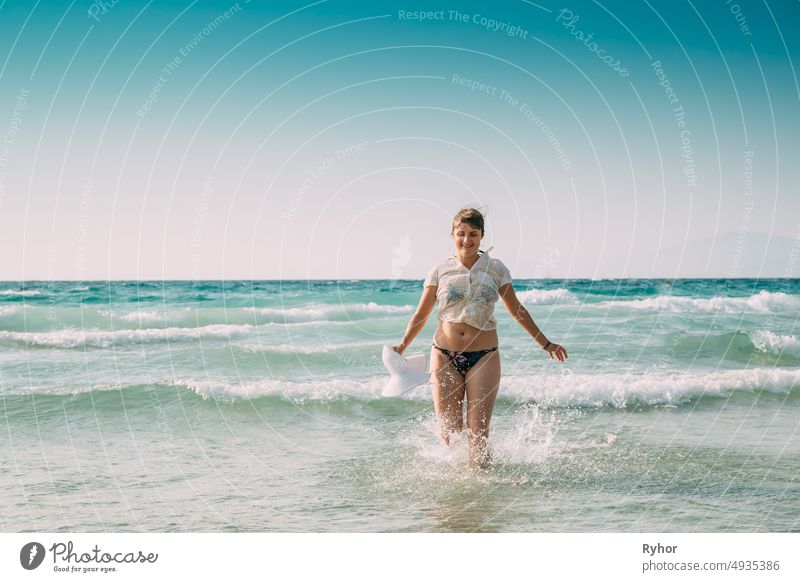 Junge kaukasische Frau Frau in Badeanzug und Sommerhut Walking in Meerwasser. Urlaub am Meer Ozean Strand Ägäis Spritzer aktiv aktiver Lebensstil attraktiv