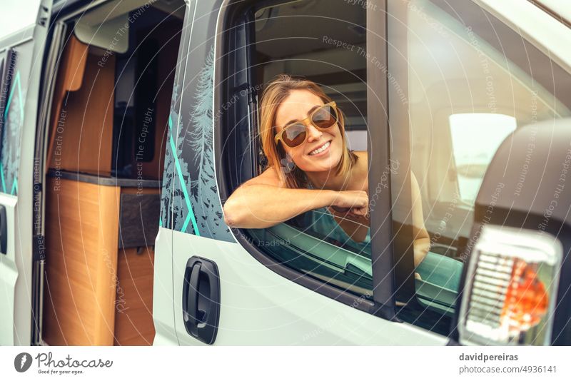 Junge Frau mit Sonnenbrille sitzt im Wohnmobil und lehnt sich an die Tür Glück jung Kleintransporter Sitzen Lehnen Sommer Ausflug reisen Lächeln Reise Fahrzeug