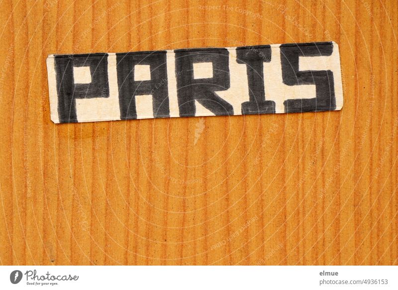 ein Pappschild mit der Aufschrift PARIS in schwarzen, handgeschriebenen Druckbuchstaben ist an eine hellbraune Holzstrukturwand geklebt Paris Zettel Handschrift