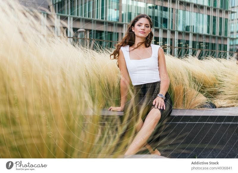 Wunderschöne junge Frau sitzt auf einer Bank in einem modernen Stadtpark selbstsicher Park Gebäude Großstadt Porträt selbstbewusst Vorschein Straße traumhaft