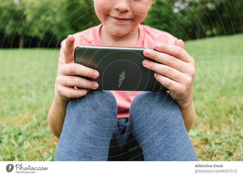 Glücklicher Junge mit Smartphone auf der Parkwiese zuschauen Video Rasen benutzend Wochenende Sommer Zeitvertreib Kind Apparatur Gerät Funktelefon Freude Gras