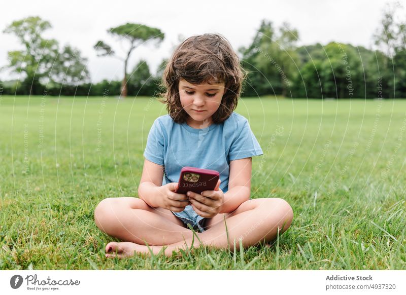 Glückliches Mädchen mit Smartphone auf einer Parkwiese zuschauen Video Rasen benutzend Wochenende Sommer Zeitvertreib Kind Apparatur Gerät Funktelefon Freude