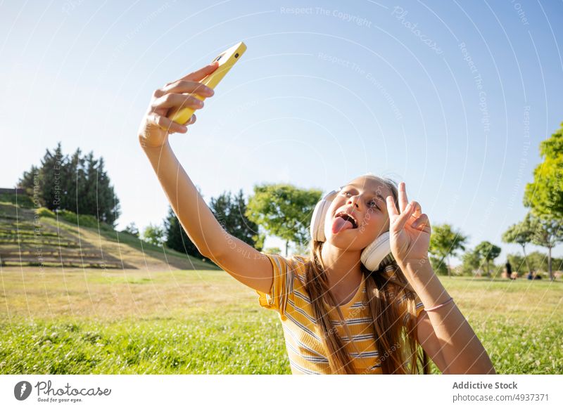 Fröhliches Mädchen mit Kopfhörern macht Selfie im Park Teenager Smartphone v-Zeichen Fotografie soziale Netzwerke Musik zuhören Frieden zwei Finger