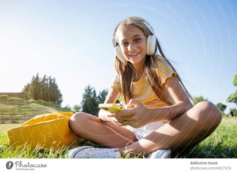 Lächelndes Mädchen mit Kopfhörern, das im Park auf seinem Smartphone surft Teenager Browsen online Musik zuhören meloman Zeitvertreib Freizeit Rasen positiv