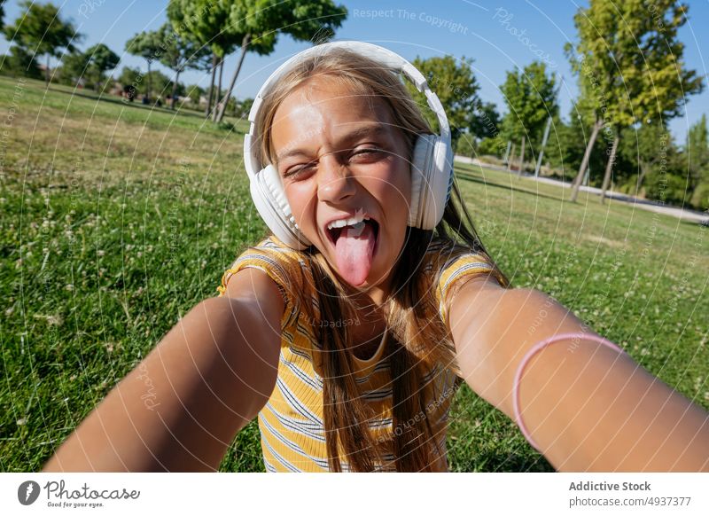 Fröhliches Mädchen mit Kopfhörern macht Selfie im Park Teenager Smartphone Fotografie soziale Netzwerke Musik zuhören gestikulieren ausspannen Zunge zeigen