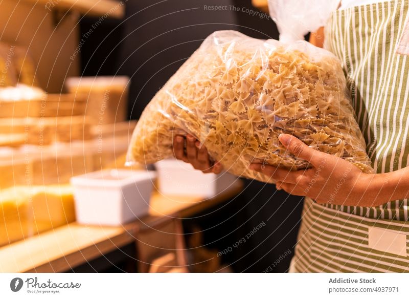 Crop-Frau mit Tüte Nudeln Spätzle Anbieter Tasche führen Arbeit Lebensmittelgeschäft Farfalle Werkstatt Uniform Schürze organisch Laden Paket Produkt Job