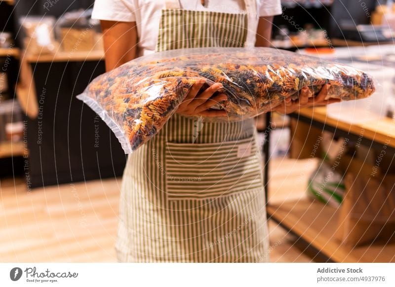 Crop-Frau mit Tüte Nudeln Spätzle Anbieter Tasche führen Arbeit Lebensmittelgeschäft Spiralnudel Werkstatt Uniform Schürze organisch Laden Paket Produkt Job