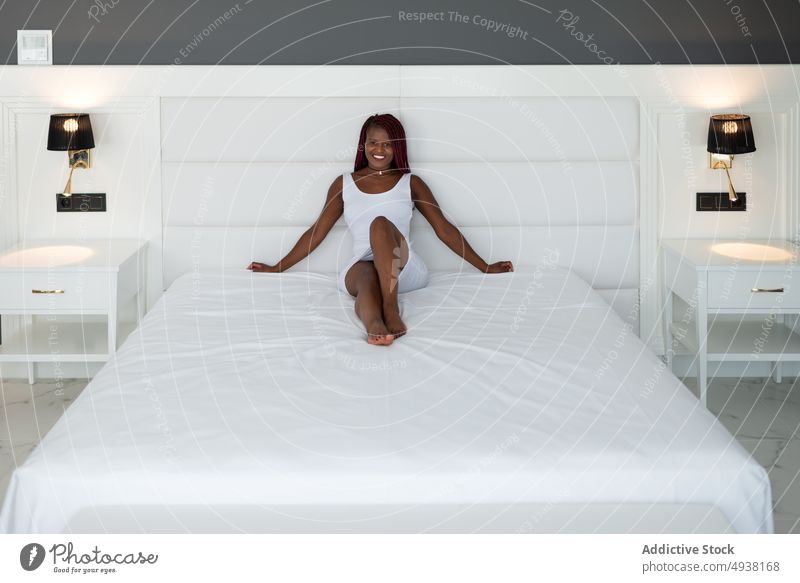 Glückliche schwarze Frau auf dem Bett sitzend Lächeln Stil Schlafzimmer heiter Barfuß Hotel Vorschein Komfort jung Afroamerikaner ethnisch weißes Kleid positiv