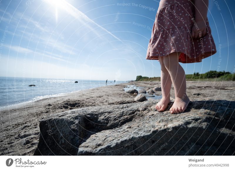Kind steht auf einem Felsen am Strand mit Blick auf das Ostsee-Meer und Dünen im Hintergrund Nordsee Sand Sandstrand barfuß Stein Norddeutschland