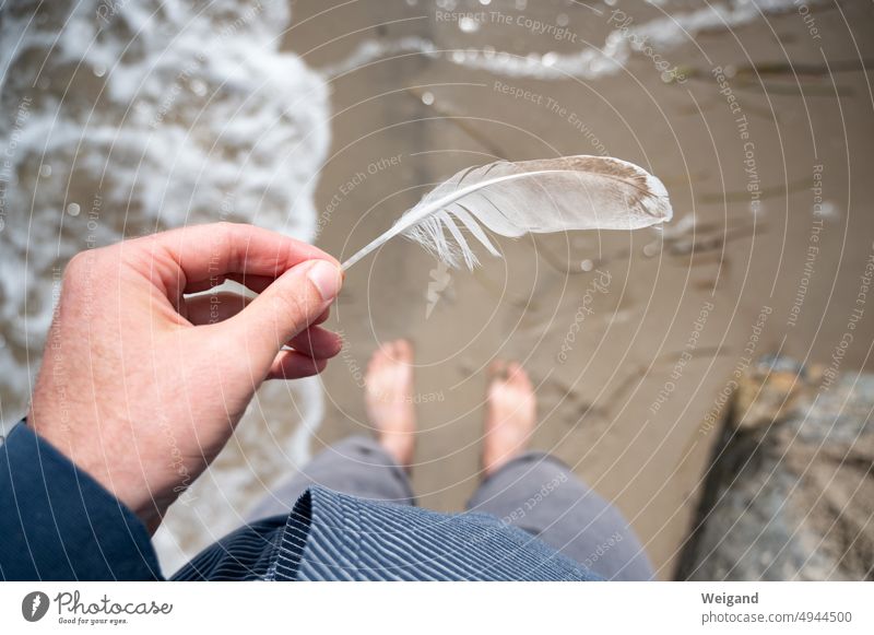 Eine weiße Feder in der Hand am Strand mit Blick nach unten auf die Füße im Wasser Achtsamkeit Urlaub Strandurlaub Ostsee Meer Nordsee Norddeutschland