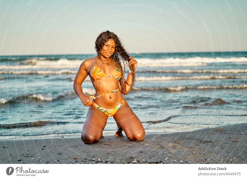 Afroamerikanische Frau kniend am Meer MEER Strand Wochenende Haare berühren Sommer Sonnenuntergang Urlaub Resort Feiertag schwarz Afroamerikaner ethnisch Bikini
