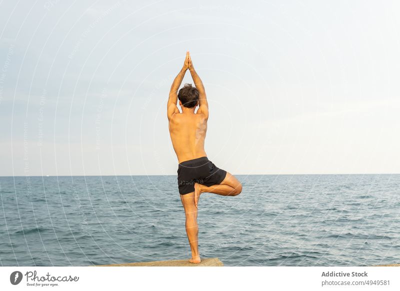 Anonymer Mann in Baum-Pose am Meer Yoga Stauanlage MEER Sitzung Baumhaltung Gleichgewicht Felsen Wasser üben männlich Stressabbau Himmel grau Zen Ufer Asana