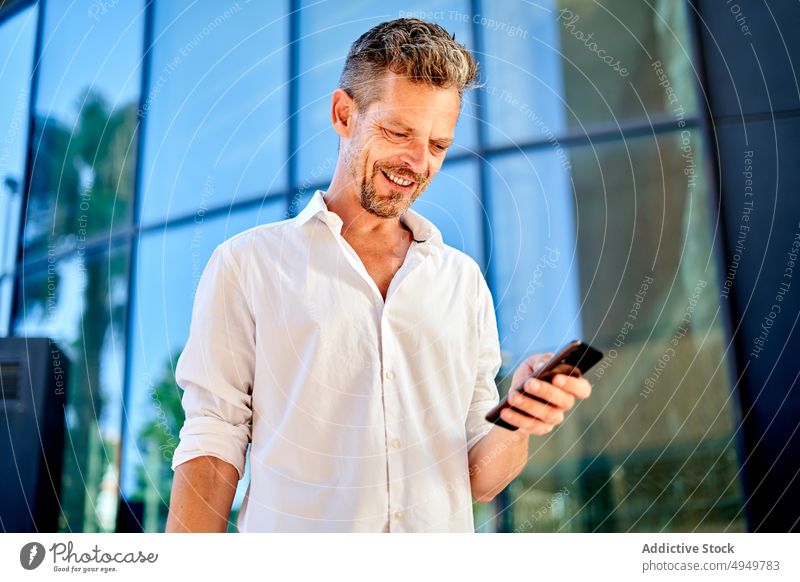 Geschäftsmann benutzt Smartphone in der Nähe einer Glaswand benutzend Straße Lächeln soziale Netzwerke Glück Manager Browsen Gebäude urban männlich Lebensmitte
