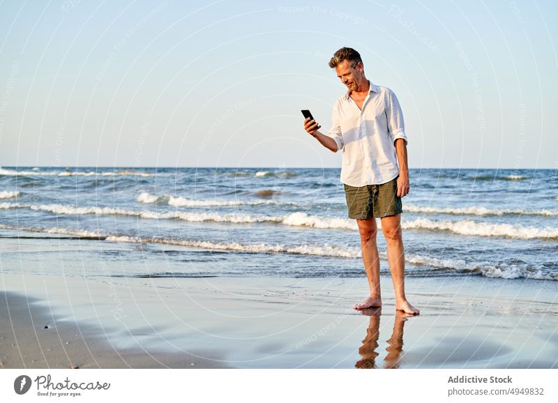 Geschäftsmann, der am Meer spazieren geht und sein Smartphone benutzt Spaziergang MEER Texten benutzend Urlaub Strand Sommer Telearbeit männlich Resort Barfuß