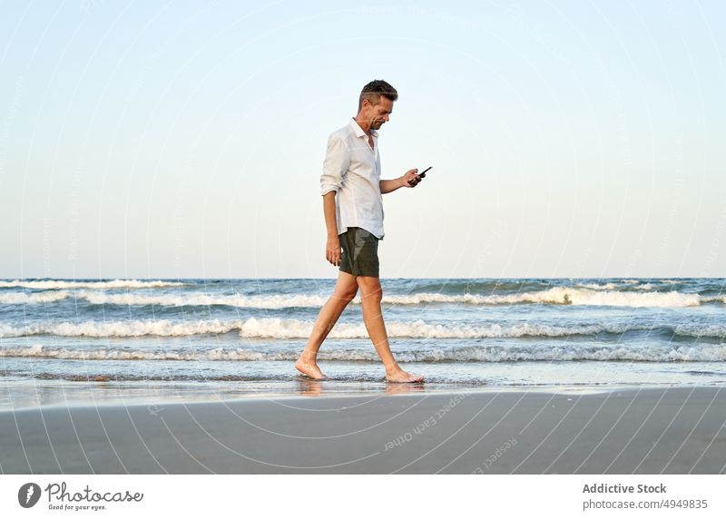 Geschäftsmann, der am Meer spazieren geht und sein Smartphone benutzt Spaziergang MEER Texten benutzend Urlaub Strand Sommer Telearbeit männlich Resort Barfuß