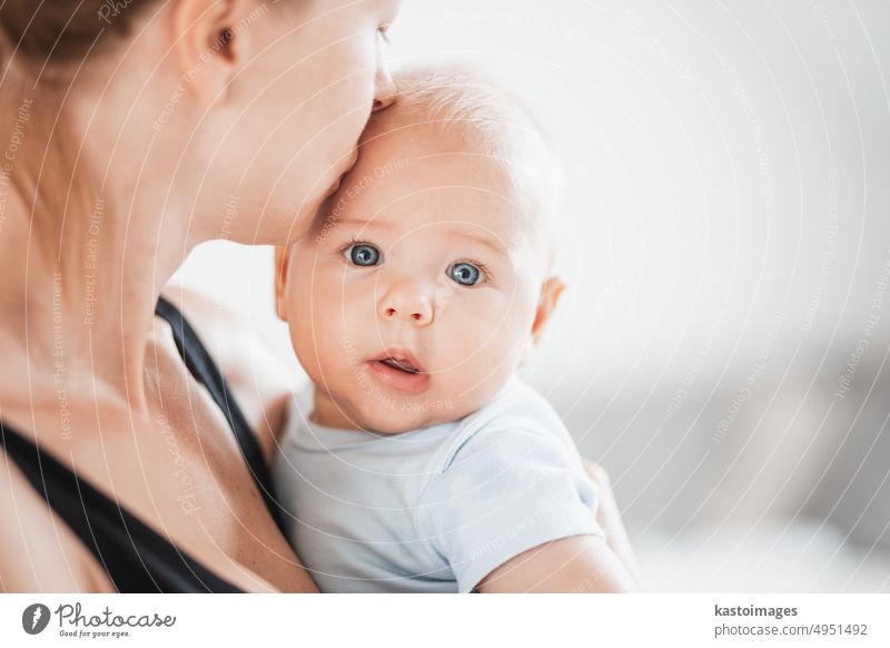Porträt eines süßen Babys, das in den Armen seiner Mutter ruht und in die Kamera schaut. Neue Mutter hält und küsst kleines Kind, umarmt Kind mit Zärtlichkeit, Liebe, Pflege. Mutterschaft Konzept.
