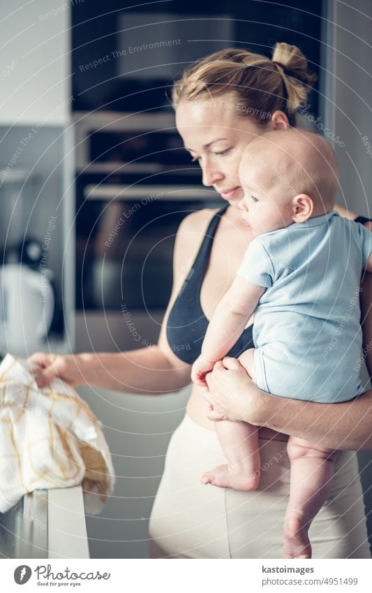 Eine Frau wischt die Spüle mit einem Tuch ab, nachdem sie den Abwasch beendet hat, während sie ein vier Monate altes Baby in ihren Händen hält. Kindheit Familie
