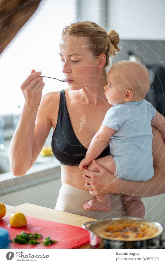 Frau kocht und tasert Essen, während sie ein vier Monate altes Baby in den Händen hält. Kindheit Familie Mutterschaft Eltern Küche heimwärts Sohn Junge
