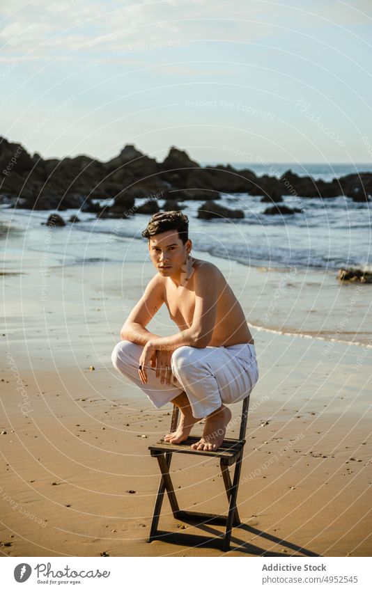 Hemdloser Mann sitzt auf Knien auf einem Stuhl am Meer Strand MEER Stil stehen Gleichgewicht Konzept verwundbar Umarmen männlich Model jung ohne Hemd Barfuß