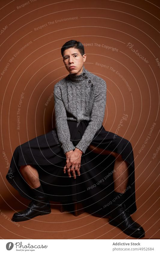 Stilvolles homosexuelles Modell im Pullover auf braunem Hintergrund schwul Hände gefaltet feminin lgbt Mann emotionslos Porträt gestrickt Stoff Stiefel Leder