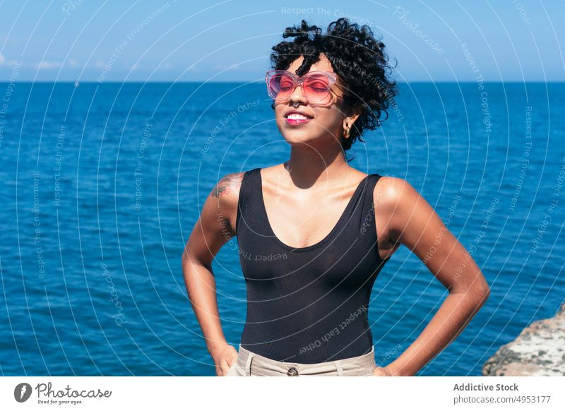 Lächelnde schwarze Frau mit Sonnenbrille vor endlosem Meer heiter Individualität Stil Tattoo feminin Hand auf der Taille MEER Blauer Himmel modern kreativ